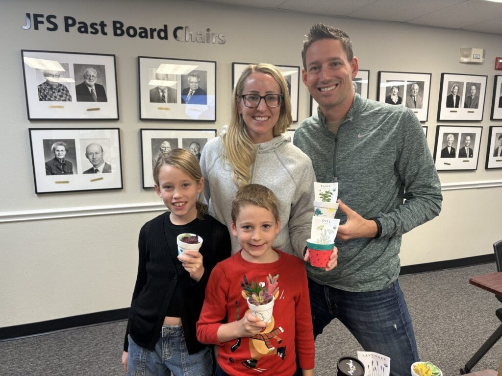 A smiling family of four holds potted plants in a room with photos of past board chairs on the wall behind them.