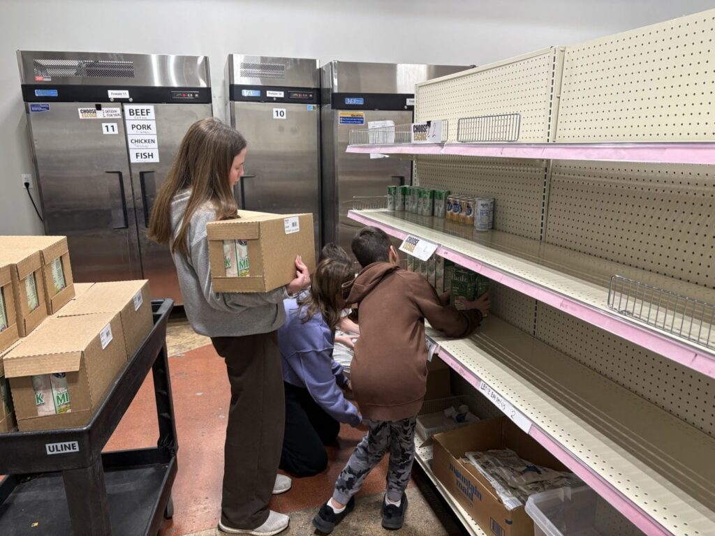 Four people restock canned goods on nearly empty shelves in a storage room, with boxes and carts nearby.