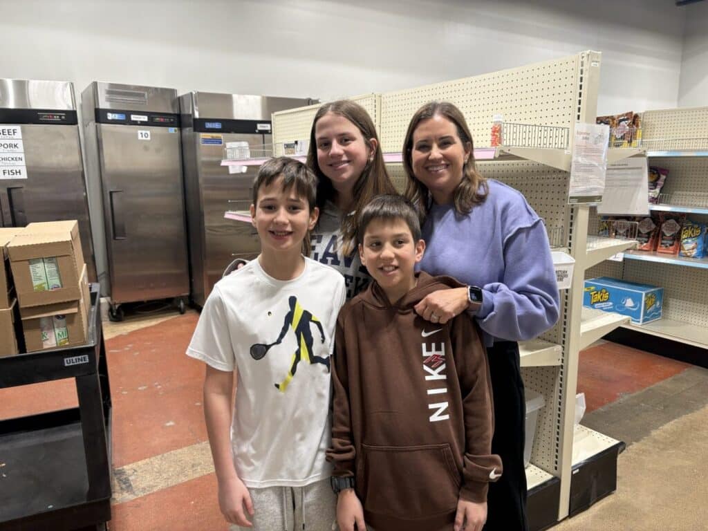 A woman and three kids smile together in a store aisle with shelves and refrigerators in the background.