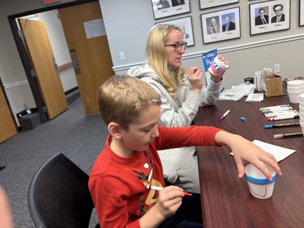 A boy and a woman decorate white cups with markers at a table in a room with framed photos on the wall.