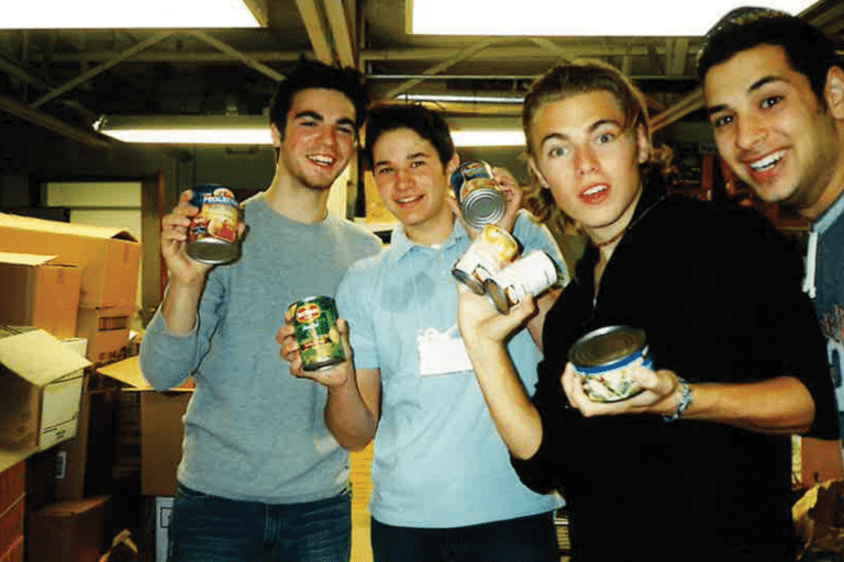 Four smiling young men stand indoors holding canned goods, with boxes stacked in the background.