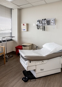 A clean, empty doctor's exam room with a medical table, chair, equipment on the wall, and window blinds.