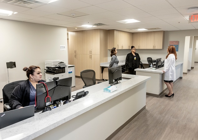 Reception area of a medical office with staff at desks and two people talking near the back counter.