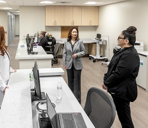 Three women stand talking in a modern medical office, with staff working at computers in the background.