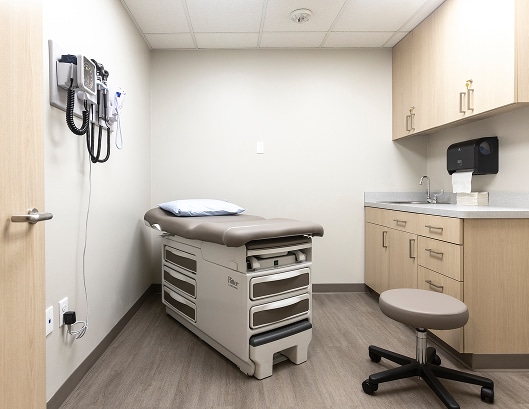 A clean, empty medical exam room with an exam table, stool, cabinets, and medical equipment on the wall.