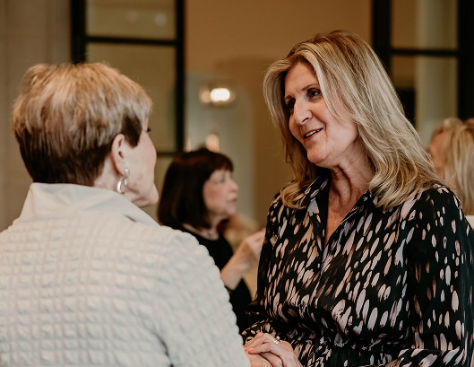 Two women are engaged in conversation at an indoor social event, with others talking in the background.