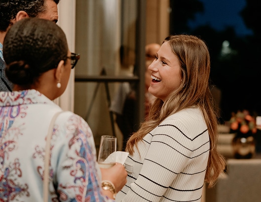 A woman in a striped top smiles and laughs while chatting with two people at an indoor event.