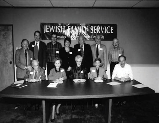 A group of 12 people pose at a table under a "Jewish Family Service" banner in a meeting room.