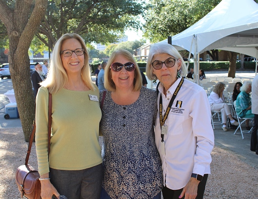 Three women smiling and posing together outdoors at a sunny event with tents and people in the background.