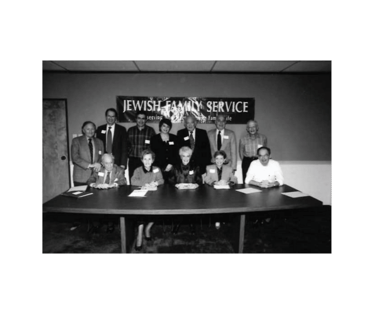 Black and white photo of thirteen people at a table under a "Jewish Family Service" banner, posing for a group picture.
