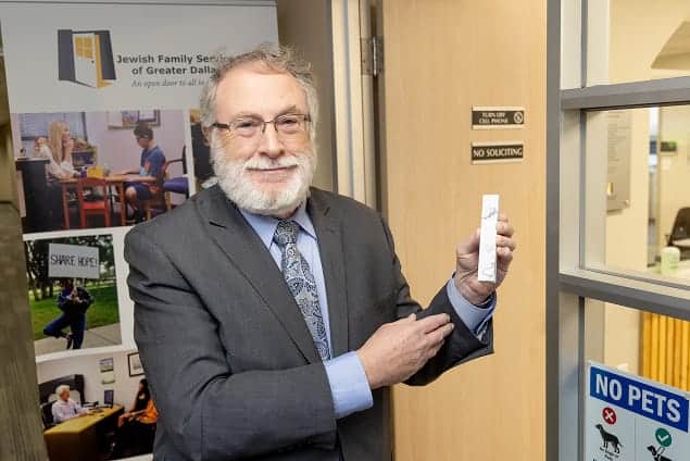 Smiling man in a suit holds a small box and points to it, standing by an office door with informational signs.