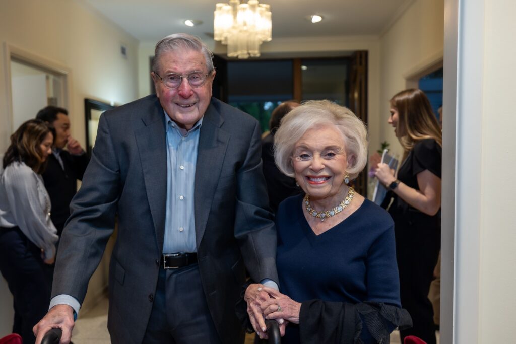 An elderly man and woman dressed formally, smiling and holding hands at an indoor gathering with others in the background.