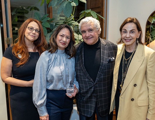 Four adults stand closely together indoors, smiling at the camera, with plants and a mirror visible in the background.