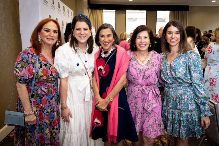 Five women smiling and posing together at an indoor event, all wearing colorful, patterned dresses.