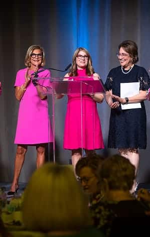 Three women stand on stage, two in pink dresses and one in black, speaking at a clear podium during an event.