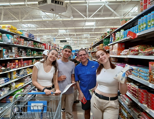 Four people smiling in a grocery store aisle, with shelves of food and a shopping cart in the foreground.