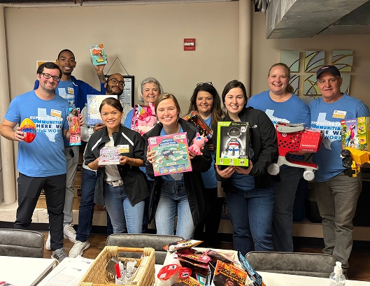 A group of smiling people holds up toys in a room, wearing blue shirts and name tags, standing behind a table.