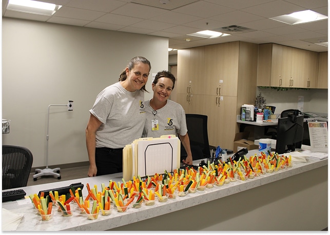 Two women in gray shirts stand behind a counter covered with trays of colorful cut vegetables in an office setting.