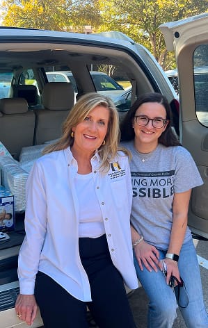 Two women smiling while sitting on the back of an open SUV, with boxes and supplies visible inside the vehicle.