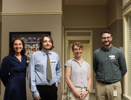 Four adults stand smiling in a hallway, dressed in business casual attire, with a painting on the wall behind them.