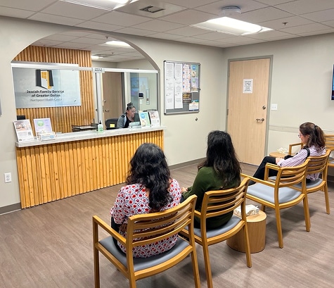 Three women sit in a waiting room with wooden chairs, facing a reception desk where a staff member is seated.