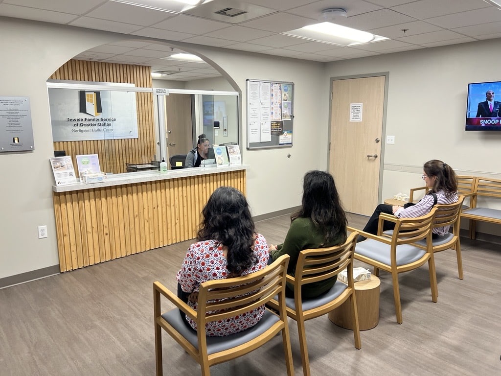 Three women sit in a waiting room with wooden chairs, facing a reception desk where a staff member is working.