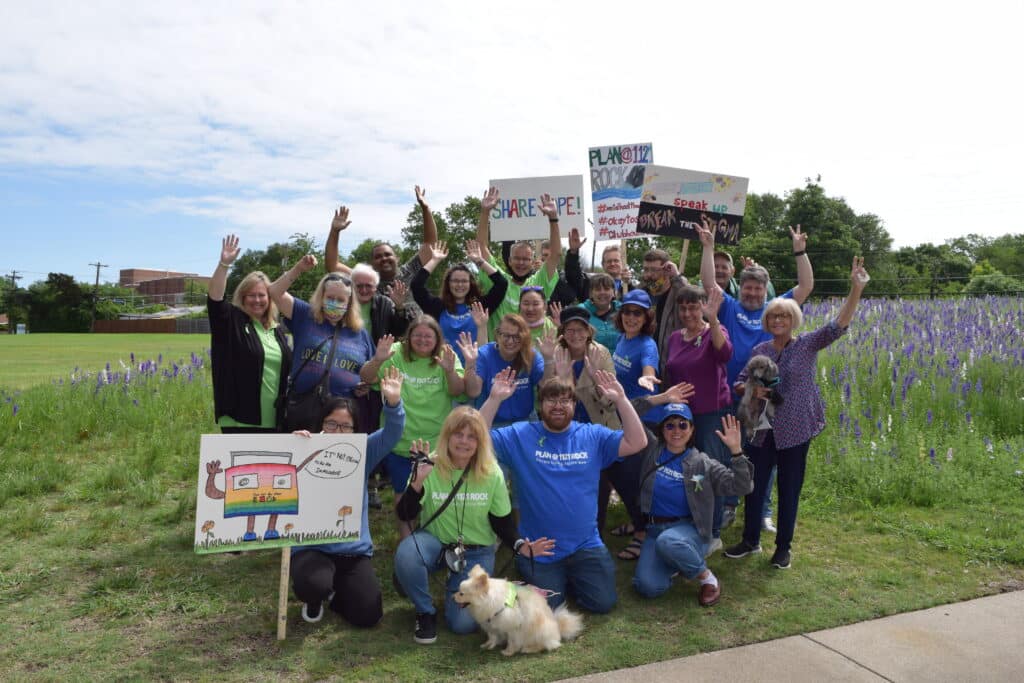 A diverse group cheers outdoors, holding signs and dogs, with wildflowers and greenery in the background.