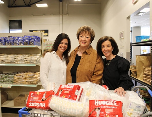 Three women stand smiling behind a cart filled with Huggies diapers in a store with shelves of packaged food.