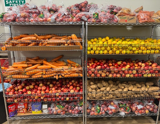 Shelves stocked with carrots, apples, potatoes, lemons, onions, and packaged foods in a market or storage area.