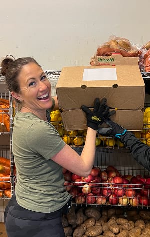 Smiling woman lifts a box of produce in front of shelves filled with fruits and vegetables.