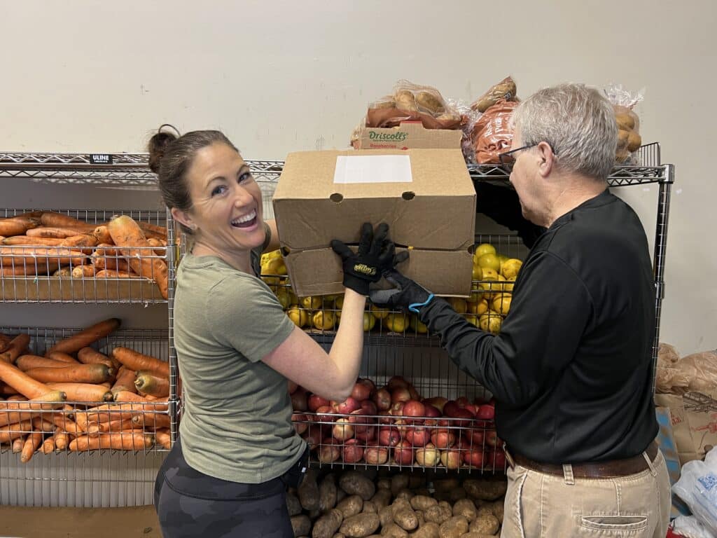 Two people smiling and lifting a box in front of shelves filled with carrots, potatoes, apples, and other produce.