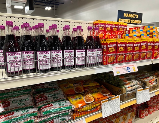 Bottles of Kedem grape juice and boxes of mac & cheese on a shelf in a kosher food section at a grocery store.