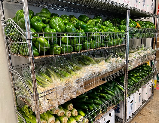 Metal shelves stacked with green bell peppers, packaged green onions, and cucumbers in a grocery or storage area.