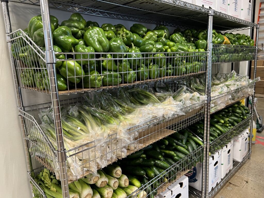 Metal shelves filled with green bell peppers, packaged green onions, and cucumbers in a fresh produce storage area.