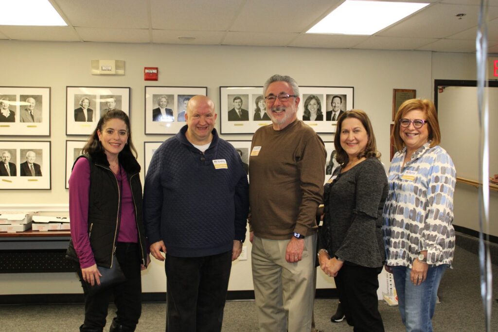 Five people stand smiling in an office with framed portraits on the wall behind them.