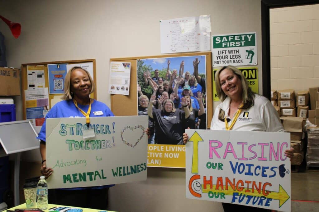 Two women smile and hold colorful signs promoting mental wellness and positive change in an office setting.