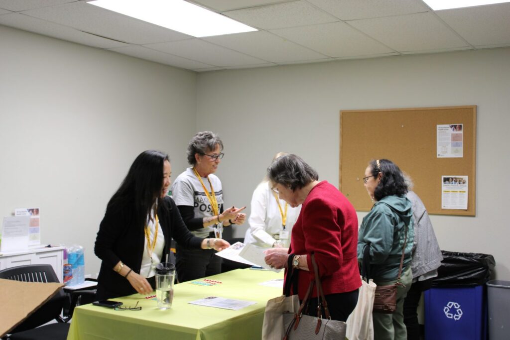 Four women stand at a table with papers in a brightly lit room, engaging in conversation near a corkboard and recycling bin.