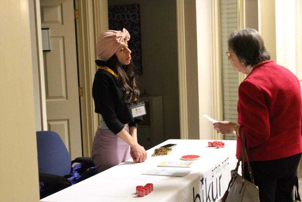 A young woman at a registration table speaks with an older woman in a red jacket in a hallway.
