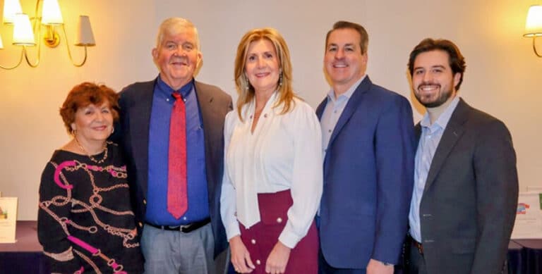 Five adults dressed in business attire stand together smiling in a well-lit room with a light-colored wall and sconces.
