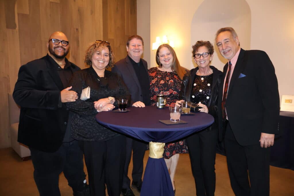 Six people dressed in formal attire stand around a cocktail table, smiling at the camera at an indoor event.