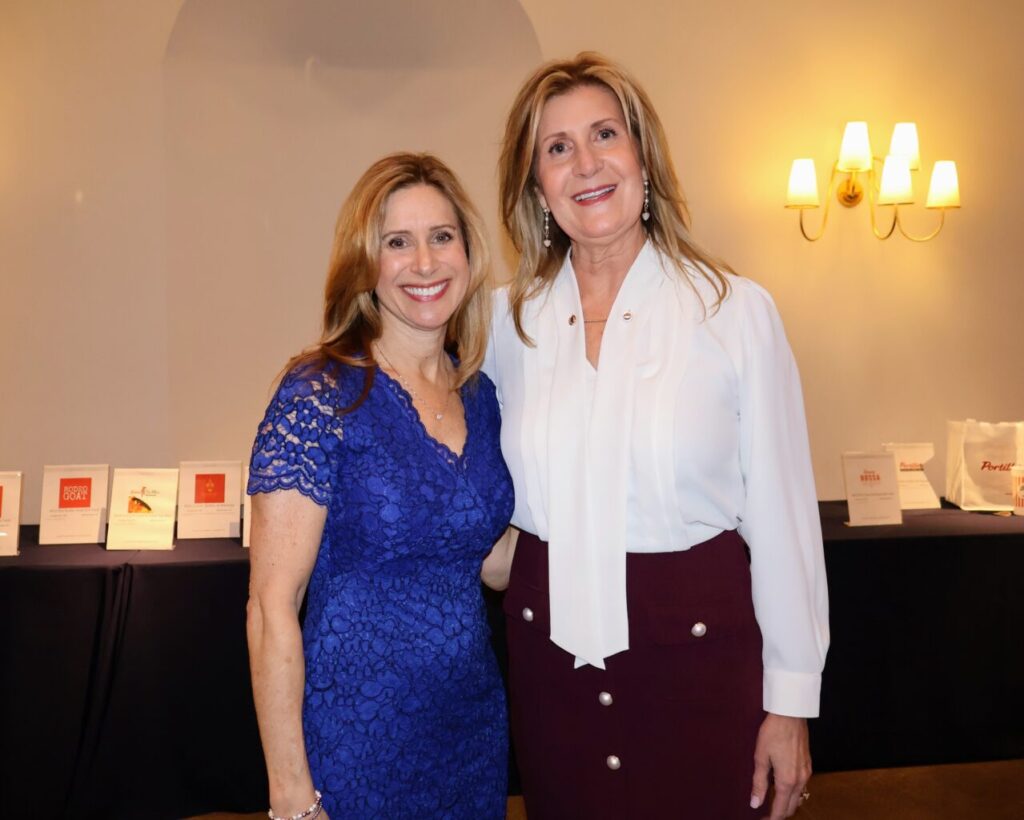Two women smiling and posing together indoors, with a table and wall lights in the background.