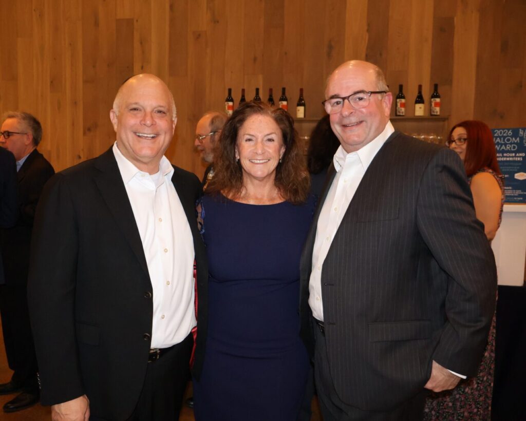 Three people in formal attire smile and pose together at an indoor event with wooden walls and bottles on a shelf behind them.