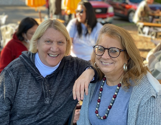 Two women smiling outdoors, one wearing glasses and a beaded necklace, with people and cars in the background.