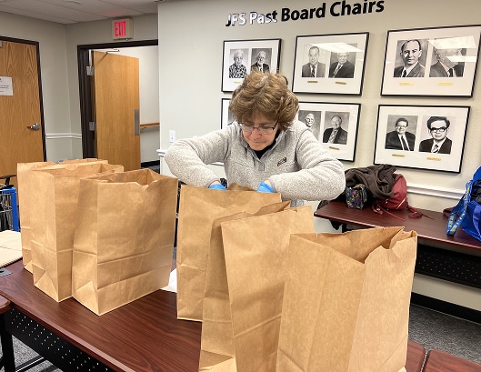 A woman fills brown paper bags at a table in an office, with portraits of past board chairs on the wall behind her.