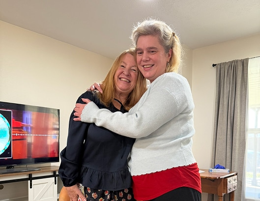 Two women smiling and hugging each other in a living room with a TV and window in the background.
