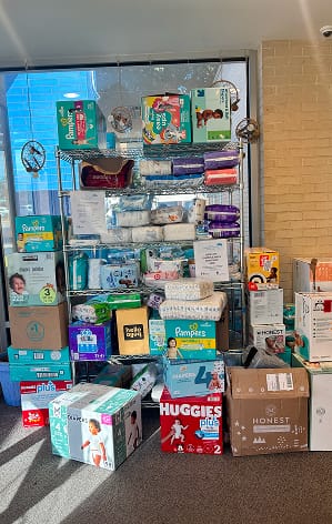 Shelves and floor stacked with various brands of diapers and wipes in a donation or collection area by a window.