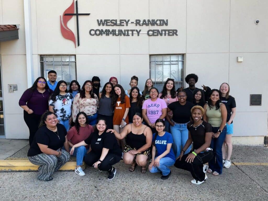 A diverse group of people smiles and poses together outside the Wesley-Rankin Community Center building.
