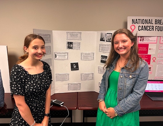 Two young women stand smiling in front of a project display board at a presentation event.