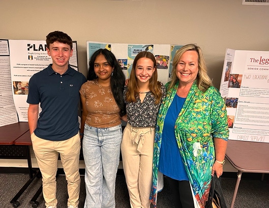 Four people stand smiling in front of project display boards in a classroom or event setting.