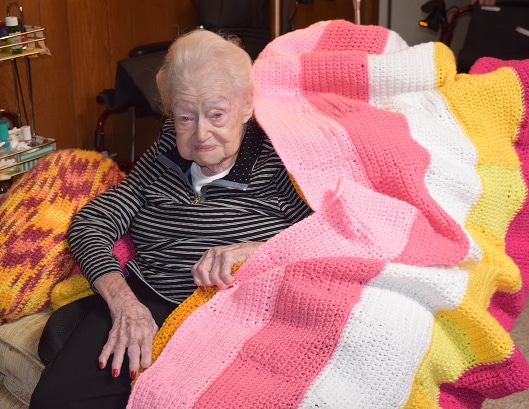 Elderly woman sitting on a couch, covered with a colorful striped knit blanket in pink, white, and yellow.